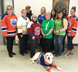 The Bonnyville Victim Services staff posing sport jerseys, Odie the support dog in front with a jersey on.